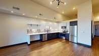 This thoughtfully designed single-floor unit showcases a contemporary aesthetic with high vaulted ceilings that lend an open, airy quality throughout. The kitchen is a standout feature, boasting a striking two-tone cabinet scheme with crisp white upper cabinets and deep navy lower cabinets, complemented by a bold decorative patterned tile backsplash. White quartz countertops, a stainless steel undermount sink, and a full suite of stainless steel appliances — including a refrigerator, range, microwave, and dishwasher — make this kitchen as functional as it is visually appealing. Open floating shelves add a modern touch alongside ample upper cabinet storage. The living area is bathed in natural light from a floor-to-ceiling multi-panel window and door system, offering expansive views of the surrounding mountains and cityscape. Rich wood-tone flooring flows consistently throughout the space, anchored by a sleek multi-arm chandelier overhead. The bathroom features dark cabinetry with gold hardware accents, wood-tone flooring, and clean white walls for a refined, modern finish. A dedicated utility area houses a stacked washer and dryer unit alongside a water heater, providing practical in-unit convenience. Dark-framed interior doors with matte black and gold hardware complete the cohesive, modern design language found throughout.