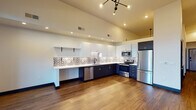This thoughtfully designed single-floor unit showcases a contemporary aesthetic with high vaulted ceilings that lend an open, airy quality throughout. The kitchen is a standout feature, boasting a striking two-tone cabinet scheme with crisp white upper cabinets and deep navy lower cabinets, complemented by a bold decorative patterned tile backsplash. White quartz countertops, a stainless steel undermount sink, and a full suite of stainless steel appliances — including a refrigerator, range, microwave, and dishwasher — make this kitchen as functional as it is visually appealing. Open floating shelves add a modern touch alongside ample upper cabinet storage. The living area is bathed in natural light from a floor-to-ceiling multi-panel window and door system, offering expansive views of the surrounding mountains and cityscape. Rich wood-tone flooring flows consistently throughout the space, anchored by a sleek multi-arm chandelier overhead. The bathroom features dark cabinetry with gold hardware accents, wood-tone flooring, and clean white walls for a refined, modern finish. A dedicated utility area houses a stacked washer and dryer unit alongside a water heater, providing practical in-unit convenience. Dark-framed interior doors with matte black and gold hardware complete the cohesive, modern design language found throughout.