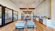 Located at 13000 Saginaw Blvd in Fort Worth, Texas, this thoughtfully designed single-story space blends modern farmhouse aesthetics with contemporary finishes to create a welcoming and functional environment. The open-concept layout is anchored by soaring ceilings adorned with rich, dark exposed wood beams that add warmth and architectural character throughout. Wide-plank hardwood-style flooring runs the length of the space, tying together the various functional zones seamlessly. The main living area is bathed in natural light, courtesy of expansive floor-to-ceiling windows and black-framed double glass entry doors with transom windows above, offering views to the outdoors. A statement chandelier with gold accents and elongated glass pendants serves as a striking focal point overhead. A sage green shiplap accent wall with an inset flat-screen television anchors one end of the living area, complemented by neutral, contemporary furnishings arranged for comfort and conversation. A built-in refreshment station with white cabinetry, a beverage refrigerator, and a tiled backsplash provides convenient amenity service. Black-framed sliding barn doors offer separation to additional rooms, while multiple seating vignettes and an adjoining dining area complete this versatile and inviting space.