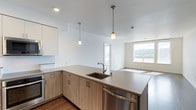 This contemporary space showcases a thoughtfully designed open-concept layout, merging the kitchen and living areas for a spacious, airy feel. The kitchen features sleek, light wood cabinetry with minimalist hardware, creating a clean and modern aesthetic. A expansive countertop with an undermount sink and stylish faucet provides ample workspace. Stainless steel appliances, including a dishwasher, add a professional touch to the culinary space. Overhead, two pendant lights with frosted glass shades offer focused illumination. The smooth transition to the living area is marked by hardwood flooring that runs throughout. A large window in the adjacent space floods the area with natural light, enhancing the bright and open atmosphere. The crisp white walls and ceiling contribute to the overall sense of spaciousness and modernity in this well-designed, functional living space.