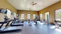 Located at 501 Trade Wind Place in Chesapeake, Virginia, this single-level fitness center offers a comprehensive workout environment designed to accommodate a wide range of exercise routines. The facility is divided into distinct functional zones, each outfitted with purpose-specific equipment.

The primary cardio area is generously sized, featuring treadmills, elliptical trainers, stationary bikes, and rowing machines arranged across durable, dark rubber flooring. Warm golden-tan walls, wood-trimmed windows, and recessed ceiling lighting create an inviting atmosphere, while dual ceiling fans with wood-toned blades promote airflow throughout the space. Multiple wall-mounted televisions are positioned throughout for entertainment during workouts.

Large windows line the walls on multiple sides, flooding the space with natural light and offering views of the surrounding outdoor environment. Full-length mirrors along one wall visually expand the room and allow users to monitor their form during exercise.

A separate strength and free weights area features a hardwood floor, floor-to-ceiling mirrors, stability balls, adjustable benches, and additional cable machines, providing a dedicated space for resistance training and functional fitness. The overall design balances practicality with a polished aesthetic, making this fitness center a well-rounded facility for diverse training needs.