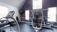 This dedicated fitness room offers a well-equipped workout environment characterized by a striking vaulted ceiling that lends a sense of openness to the space. The bold purple accent walls create an energizing atmosphere, contrasting with crisp white trim and a neutral blue-gray rubberized flooring ideal for exercise use. Natural light fills the room through two windows, including a large upper window positioned beneath the peak of the vaulted ceiling, brightening the space during daytime workouts. A wall-mounted flat-screen television is positioned for convenient viewing during cardio sessions. The room is outfitted with a multi-station cable weight machine that supports a variety of strength training exercises, as well as a treadmill and a stationary bike, providing options for both cardiovascular and resistance training. A small shelf unit holds additional equipment or accessories near the cardio area. The combination of functional flooring, ample equipment, and thoughtful layout makes this space a practical and motivating environment for personal fitness routines.