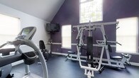 This dedicated fitness room offers a well-equipped workout environment characterized by a striking vaulted ceiling that lends a sense of openness to the space. The bold purple accent walls create an energizing atmosphere, contrasting with crisp white trim and a neutral blue-gray rubberized flooring ideal for exercise use. Natural light fills the room through two windows, including a large upper window positioned beneath the peak of the vaulted ceiling, brightening the space during daytime workouts. A wall-mounted flat-screen television is positioned for convenient viewing during cardio sessions. The room is outfitted with a multi-station cable weight machine that supports a variety of strength training exercises, as well as a treadmill and a stationary bike, providing options for both cardiovascular and resistance training. A small shelf unit holds additional equipment or accessories near the cardio area. The combination of functional flooring, ample equipment, and thoughtful layout makes this space a practical and motivating environment for personal fitness routines.