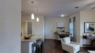 This single-floor residence showcases an open-concept layout that seamlessly connects the kitchen and dining areas. The kitchen features a granite countertop island with a built-in sink and bar seating for two, complemented by sleek pendant lighting that hangs above. White cabinetry and modern fixtures contribute to a clean, contemporary aesthetic throughout the space.

The dining area is positioned adjacent to the kitchen and centers around a round dark-wood table surrounded by upholstered chairs. A framed abstract artwork adorns the wall nearby, adding a decorative focal point to the space. Natural light filters in through a window dressed with neutral-toned blinds, brightening the area.

Dark hardwood flooring runs throughout, providing a rich contrast to the white walls and light-colored furnishings. The entryway features a white front door, with an interior door visible toward the rear, suggesting additional private rooms. The overall ambiance is modern and cohesive, with a neutral color palette and thoughtful lighting creating a welcoming, well-appointed atmosphere.