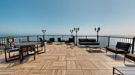 Located at 1400 N Lake Shore Drive in Chicago's prestigious Gold Coast neighborhood, this rooftop terrace offers an exceptional outdoor amenity space with sweeping views of Lake Michigan and the surrounding cityscape. The terrace is finished with elegant wood-look porcelain tile decking laid in a herringbone pattern, lending a refined, contemporary aesthetic underfoot. Raised stepped sections add architectural dimension to the layout, while classic multi-arm black lantern-style lamp posts are positioned throughout, providing both ambient lighting and a sophisticated design accent. A sturdy black metal railing frames the perimeter, ensuring safety while preserving unobstructed sightlines toward the lake and open sky. The space is thoughtfully furnished with multiple seating arrangements, including a sofa, lounge chairs, and side tables, creating inviting areas for relaxation and social gatherings. A green living wall panel introduces a natural, lush element to the hardscape environment. Gas grills are available on-site, making the terrace well-suited for outdoor entertaining. Access is provided through a frosted glass door framed by richly textured slate tile walls. Signage throughout reflects the building's managed, shared-use nature, underscoring the terrace's role as a curated communal amenity.