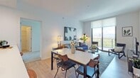 This contemporary single-floor residence is designed with a clean, modern aesthetic and an open-concept layout that seamlessly connects the living, dining, and kitchen areas. Light wood-tone flooring flows throughout the main living spaces, complementing the bright white walls and recessed lighting overhead.

The living area is anchored by floor-to-ceiling windows that flood the space with natural light and offer expansive views of the surrounding cityscape and sky. A glass-top coffee table and stylish seating arrangements make this an inviting area for relaxation and entertaining.

The dining area features a marble-topped table with mixed seating, positioned to take advantage of the natural light and views. The kitchen is outfitted with crisp white cabinetry, a large quartz waterfall island with bar seating for multiple guests, and a full suite of stainless steel appliances including a refrigerator, microwave, and range. A stacked washer and dryer unit is conveniently tucked adjacent to the kitchen.

The primary bedroom offers plush carpeting, framed wall art, and direct access to an en-suite bathroom, along with a dedicated closet space. The second bedroom is equally well-appointed, featuring soft carpeting and access to a full bathroom complete with a soaking tub and shower combination. Throughout, the space maintains a cohesive, contemporary feel with thoughtful finishes and functional design.