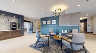 This single-floor lobby and amenity space for Aura Luxury Living combines contemporary design with welcoming warmth. The entrance features expansive floor-to-ceiling glass doors that flood the space with natural light, opening onto large-format light tile flooring that flows throughout. A dramatic focal point anchors the lounge area: a tiled fireplace clad in stacked blue-grey stone, topped with a chevron-patterned wood panel surround and framed by a deep teal accent wall displaying a curated trio of framed artwork. Mid-century modern lounge chairs in soft grey upholstery are arranged around a round live-edge wood coffee table, all set atop a bold navy patterned area rug, creating an inviting seating vignette. An eye-catching multi-arm chandelier with black and brass accents provides warm overhead illumination. Adjacent to the main lounge, a built-in credenza with a patterned tile backsplash serves as a coffee and refreshment station. The space extends further into additional amenity rooms, including what appears to be a resident lounge or co-working area visible through glass-paneled doors. Recessed lighting and wide corridors connect the various areas, maintaining an open, airy ambiance throughout the well-appointed communal space.