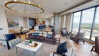 Located at 1007 Church St in Evanston, Illinois, this elevated single-floor space delivers a refined, contemporary design with an open, flowing layout. Light oak hardwood flooring extends throughout, unifying the various functional zones beneath crisp white ceilings with recessed lighting.

The expansive lounge area serves as the centerpiece, anchored by a statement circular brass chandelier and a wall-mounted display above a sleek fireplace. Multiple seating arrangements — including plush sofas, accent chairs, and side tables — offer flexible social configurations. Exposed concrete columns add an industrial counterpoint to the otherwise polished interior.

Floor-to-ceiling windows wrap around the perimeter, flooding the space with natural light and framing panoramic views of the Evanston skyline and Lake Michigan beyond. Sheer linen drapery softens the glass line while preserving the visual connection to the outdoors.

A fully equipped kitchen and bar area features dark cabinetry, stainless steel appliances, a textured metallic backsplash, and bar seating, making it well-suited for catered events or casual gatherings. An adjacent dining nook showcases a round marble-topped table paired with upholstered chairs and a colorful landscape painting.

Curated artwork displayed throughout the space adds warmth and personality, complementing the neutral, modern palette and creating an inviting, gallery-like atmosphere.