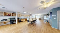 Located at 650 S San Ildefonso Rd in Los Alamos, New Mexico, this single-floor community space offers a well-organized and practical layout designed for shared use. Light hardwood flooring runs throughout, lending warmth and visual continuity across all areas.

The kitchen is efficiently designed with light maple cabinetry, a gas range, over-the-range microwave, full-size refrigerator, and ample counter space for meal preparation. Adjacent to the kitchen, a large communal dining table with seating for multiple occupants serves as a natural gathering point, seamlessly connecting the kitchen to the broader open area.

The living room area is bright and welcoming, benefiting from generous natural light through large windows fitted with vertical blinds. Comfortable seating, including a sofa and accent chair, is arranged around a central coffee table, with New Mexico-themed artwork adorning the walls. A ceiling fan adds comfort to the space.

The laundry room is outfitted with multiple commercial-grade front-loading washers and dryers, open shelving for supplies, and a glass-panel exterior door providing additional natural light and outdoor access. The room's bold accent wall adds a distinctive visual element.

Overall, the space presents a clean, functional, and community-oriented environment suited for shared residential or facility use.