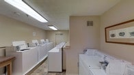 This shared laundry facility is a well-organized, single-floor space designed to accommodate the laundry needs of multiple users. The room is laid out efficiently, with rows of top-loading washing machines and dryers positioned along the walls, maximizing the use of the available floor space. A central folding counter runs along one side, providing a convenient surface for sorting and folding laundry. The walls are finished in a neutral beige tone, contributing to a clean and utilitarian atmosphere. Overhead fluorescent lighting fixtures illuminate the entire space brightly, ensuring excellent visibility throughout. The flooring consists of durable, easy-to-clean tile, well-suited to the demands of a high-traffic laundry environment. A ventilation grille is visible on the wall, supporting proper airflow within the room. A standard door at the far end of the space provides access to an adjoining area or exit. The textured ceiling adds a subtle architectural detail to an otherwise straightforward and practical space. Overall, this laundry facility is thoughtfully arranged to deliver a convenient, no-frills experience focused on functionality and ease of use.