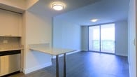 This single-floor residence showcases a clean, modern design with an open-concept living area that maximizes the sense of space and flow. The living space is anchored by full-height sliding glass doors that flood the interior with abundant natural light and provide direct access to a private balcony, offering pleasant outdoor views of surrounding greenery.

The flooring throughout is a rich, dark hardwood that contrasts elegantly with the crisp white walls and ceilings, creating a contemporary and polished aesthetic. Flush-mount ceiling light fixtures provide consistent, even illumination throughout the open area.

A kitchen peninsula with a granite-style countertop serves as a natural divider between the kitchen and living areas, offering both additional workspace and a casual seating or dining surface. The countertop's light stone finish complements the overall neutral color palette of the space.

The layout is thoughtfully designed to promote an airy, unobstructed feel, with clean sightlines from the kitchen through to the living area and out toward the balcony. The combination of natural light, modern finishes, and an open floor plan gives the space a bright, welcoming, and highly functional character suited to contemporary living.