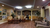 This single-floor common lounge area offers a warm and inviting atmosphere designed for comfort and community. The space features rich hardwood flooring throughout, complemented by neutral earth-toned walls adorned with decorative artwork and framed prints. A tray ceiling with recessed lighting and flush-mount ceiling fixtures provides bright, even illumination across the room.

The focal point of the space is a built-in wall unit featuring an arched alcove flanked by open display shelves and lower cabinetry finished in a warm wood tone. The shelves house books, decorative items, and a small television, making the area functional as both a library nook and media corner. A decorative medallion adds an ornamental touch to the central arch.

Seating includes wicker accent chairs with floral cushions, upholstered lounge chairs, and a sofa, all arranged around a dark wood coffee table to encourage conversation and relaxation. Potted greenery throughout adds life and warmth to the environment. The overall ambiance is homey and comfortable, reflecting a thoughtfully designed communal living space suited for everyday leisure and gathering.