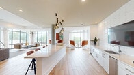 Located at 1777 Peachtree St NW in Atlanta, GA, this thoughtfully designed single-floor space offers a generous, open-plan layout that seamlessly integrates a kitchen, dining room, and living room. Floor-to-ceiling windows line the perimeter, flooding the interior with abundant natural light and providing sweeping outdoor views of the surrounding Atlanta landscape. Light-toned hardwood flooring flows throughout the main areas, complemented by plush textured carpeting in the lounge zones, creating a dynamic interplay of textures underfoot. The kitchen features a long, sleek white quartz island with bar seating, white cabinetry, a stainless steel sink, and a sculptural cluster of globe pendant lights suspended overhead. A three-dimensional white tile accent wall adds an artistic focal point. The living area is anchored by a large sectional sofa and a wall-mounted flat-screen TV, while bold orange pod-style chairs arranged near the windows offer additional intimate seating. A dedicated game and entertainment zone showcases a round poker table with vibrant orange chairs, a shuffleboard table, and a large display screen set against a striking geometric-patterned accent wall. Throughout the space, curated artwork and modern sculptural lighting fixtures contribute to a cohesive, contemporary ambiance ideal for gathering and socializing.