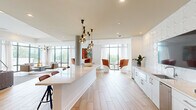 Located at 1777 Peachtree St NW in Atlanta, GA, this thoughtfully designed single-floor space offers a generous, open-plan layout that seamlessly integrates a kitchen, dining room, and living room. Floor-to-ceiling windows line the perimeter, flooding the interior with abundant natural light and providing sweeping outdoor views of the surrounding Atlanta landscape. Light-toned hardwood flooring flows throughout the main areas, complemented by plush textured carpeting in the lounge zones, creating a dynamic interplay of textures underfoot. The kitchen features a long, sleek white quartz island with bar seating, white cabinetry, a stainless steel sink, and a sculptural cluster of globe pendant lights suspended overhead. A three-dimensional white tile accent wall adds an artistic focal point. The living area is anchored by a large sectional sofa and a wall-mounted flat-screen TV, while bold orange pod-style chairs arranged near the windows offer additional intimate seating. A dedicated game and entertainment zone showcases a round poker table with vibrant orange chairs, a shuffleboard table, and a large display screen set against a striking geometric-patterned accent wall. Throughout the space, curated artwork and modern sculptural lighting fixtures contribute to a cohesive, contemporary ambiance ideal for gathering and socializing.