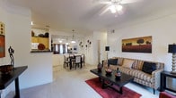 This single-floor residence offers an open-concept layout that seamlessly connects the living, dining, and kitchen areas, creating a spacious and inviting atmosphere. The living area is anchored by plush carpeting over which a bold red area rug adds a pop of color, complementing a sleek dark-wood coffee table adorned with decorative pottery. A ceiling fan with integrated lighting overhead provides both comfort and illumination. Decorative wall art and floral decals lend the space a personalized, homey character. The dining area flows naturally from the living space and features a classic dining set beneath a pendant light fixture, with a secondary ceiling fan maintaining airflow throughout. Large windows dressed with curtains allow natural light to filter in, brightening the interior. The kitchen is partially open to the living and dining areas via a raised breakfast bar counter, offering convenient pass-through access. Light wood cabinetry and a functional layout make the kitchen both practical and aesthetically cohesive. A hallway leads to additional rooms, suggesting a well-organized floor plan. The overall ambiance is warm, casual, and move-in ready, with a neutral white palette that enhances the sense of openness and light throughout the home.