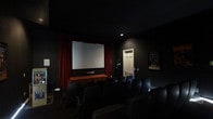 This dedicated theatre room is designed to deliver an authentic cinematic experience within a private setting. The room is finished entirely in deep black tones across the walls, ceiling, and flooring, effectively eliminating light interference and creating a fully immersive viewing environment. A large projection screen serves as the room's focal point, dramatically framed by rich red curtains that draw the eye and evoke the ambiance of a classic movie theatre. The curtains extend from ceiling to floor on either side of the screen, adding a theatrical elegance to the space. Rows of dark, cushioned recliner-style seats are arranged in a tiered layout, ensuring clear sightlines from every position in the room. A wooden console or equipment table is positioned beneath the screen, likely housing audio-visual components. A framed movie poster decorates one wall, reinforcing the cinematic theme. Subtle recessed lighting provides minimal ambient illumination without detracting from the viewing experience. A private door offers convenient access, completing this well-appointed, purpose-built theatre room.