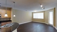 This single-floor space offers an open and airy layout designed to maximize functionality and comfort. The living area features rich, dark hardwood flooring that runs throughout, creating a cohesive and elegant foundation for the space. Warm, neutral-toned walls provide a welcoming ambiance while offering a versatile backdrop for a variety of interior styles.

The kitchen and living areas flow seamlessly into one another, defined by a curved dark granite countertop that serves as both a workspace and a visual anchor. A pendant light hangs above the counter, adding a touch of refined style and focused illumination to the kitchen zone. The living area is brightened by a combination of natural light streaming through a horizontal window fitted with blinds, as well as a ceiling-mounted flush light and an adjustable track lighting fixture that allow for flexible, layered lighting options.

Clean white baseboards and trim provide crisp contrast against the warm wall tones and dark flooring, contributing to a polished, well-finished look throughout. The open floor plan and thoughtful lighting design make this a practical and inviting space, suitable for a range of uses and furnishing arrangements.