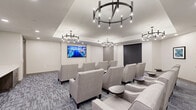 Located at 6795 Powers St in Riverdale, GA, this single-floor living room functions as a sophisticated screening and social lounge space. The room is thoughtfully designed with a contemporary aesthetic, featuring rows of plush, light gray upholstered armchairs arranged in a theater-style layout, each paired with small round side tables for added convenience. Multiple large wall-mounted flat-screen displays serve as focal points, making the space well-suited for group viewing or presentations. Overhead, a series of striking circular chandelier fixtures with exposed Edison-style bulbs hang from a recessed tray ceiling, casting warm, ambient light throughout the room. The flooring consists of a patterned commercial-grade carpet in muted gray tones that complements the neutral white and gray wall palette. A vibrant teal accent wall anchors one end of the room, adorned with colorful abstract artwork and a built-in countertop workstation with wood-toned cabinetry and a marble-look surface, adding both function and visual interest. Roller shade window treatments and taupe panel doors complete the polished, professional finish of this versatile communal space.