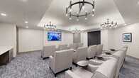 Located at 6795 Powers St in Riverdale, GA, this single-floor space comprises one thoughtfully designed living room that blends modern aesthetics with functional layout. The room accommodates multiple seating arrangements, featuring rows of light gray upholstered chairs and sofas positioned to face a large wall-mounted display screen, making it well-suited for group gatherings or presentations.

A striking teal accent wall anchors one side of the room, complemented by a built-in workstation with a marble-topped counter surface and wood-toned cabinetry beneath. Colorful framed artwork adorns the teal wall, adding a vibrant focal point. On the opposite wall, a large flat-screen display is mounted at a comfortable viewing height.

The ceiling features a recessed tray detail fitted with a contemporary ring chandelier, providing a refined overhead element alongside recessed lighting throughout. Patterned, multi-toned gray carpet covers the floor, adding texture while maintaining a neutral, cohesive palette. Taupe-painted walls keep the space feeling open and clean. Large roller shade-covered windows and multiple access doors with gray panel detailing complete the well-appointed interior.