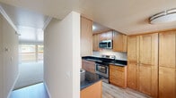 This single-floor residence offers a thoughtfully organized layout with a series of comfortable, functional spaces. The galley-style kitchen is a standout feature, boasting warm honey-toned wood cabinetry that extends to the ceiling for ample storage, rich dark granite countertops, and a full suite of stainless steel appliances including an electric range, built-in microwave, and refrigerator. Light hardwood-style flooring runs throughout the kitchen, adding a clean, modern touch. The kitchen opens toward the living area, which is generously proportioned and finished with soft wall-to-wall carpeting, crown molding, and a ceiling fan with integrated lighting. Sliding glass doors flood the living room with natural light and provide direct access to an outdoor balcony with exterior views. The bedroom continues the warm, neutral palette with carpeted flooring and features a built-in closet with mirrored sliding doors. The bathroom is clean and well-equipped, offering a white vanity with ample drawer storage, wall-mounted cabinetry, a large mirror, and a tub-shower combination with white tile surround and a glass panel enclosure. Overall, the space presents a bright, practical, and comfortable living environment.