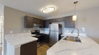 Located at 2800 John Steven Way in Reynoldsburg, Ohio, this single-floor residence offers a well-appointed layout designed for comfortable everyday living. The kitchen is a standout feature, showcasing rich dark cabinetry, striking marble-style countertops, a subway tile backsplash, and a full suite of stainless steel appliances including a refrigerator, electric range, and dishwasher. A peninsula with an integrated double sink and pendant lighting creates a natural transition into the adjoining living and dining area, which features warm hardwood-style flooring and neutral-toned walls that enhance the sense of openness. The living area flows seamlessly from the kitchen, with plush carpeting defining the lounge space and windows allowing natural light to fill the room. The bedrooms are generously sized, each finished with soft carpet, neutral walls, and windows fitted with blinds for privacy. The bathroom features a clean, modern vanity with a white countertop, a large mirror with overhead lighting, dark cabinetry below, and built-in shelving for additional storage. Throughout the space, textured ceilings and consistent neutral tones create a cohesive, move-in-ready aesthetic that balances functionality with contemporary style.