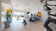 This single-level fitness center offers a bright, open workout environment designed to motivate and accommodate a wide range of exercise routines. The space features light wood-look flooring throughout, contributing to a clean, modern aesthetic complemented by whitewashed exposed brick accent walls. A Muhammad Ali quote — "Float like a butterfly, sting like a bee" — is prominently displayed on the brick wall, adding an inspirational focal point to the space.

Natural light floods the interior through floor-to-ceiling windows along one wall and a large retractable garage-style door that opens directly to an outdoor pool area, seamlessly blending indoor and outdoor environments. Ceiling fans help maintain airflow throughout the space, and mounted flat-screen televisions provide entertainment during workouts.

The fitness center is thoroughly equipped with Matrix treadmills, elliptical cross-trainers, stair climbers, and spin bikes for cardio training. A dedicated strength area includes a full rack of dumbbells, resistance bands, stability balls in multiple sizes, a yoga mat station, and a captain's chair for core work. A small workstation area is also available for members. The open layout ensures ample floor space for stretching or functional training, making this a comprehensive and well-appointed facility.