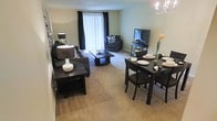 This single-floor space offers an open-concept layout that seamlessly integrates the living and dining areas into one cohesive environment. The living room is furnished with dark upholstered seating arranged around a rectangular coffee table, complemented by a side table with a decorative lamp. A flat-screen television rests on a media console positioned near a window, while an additional armchair adds extra seating. Neutral-toned carpeting runs throughout, contributing to the warm and inviting ambiance of the space.

Adjacent to the living area, the dining space features a dark wood table set for multiple guests, accompanied by matching chairs. A chandelier with candelabra-style lighting hangs above, adding an elegant touch. Soft, light-colored walls throughout the space enhance the sense of openness, while dark curtains frame a large window that allows natural light to filter in. The overall aesthetic balances contemporary dark furnishings against lighter wall tones, creating a well-defined yet connected living and dining environment suitable for both everyday living and entertaining.