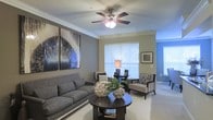 This single-story space showcases a thoughtfully designed living room that balances comfort and style. The room is anchored by a plush gray sofa adorned with accent pillows, complemented by a pair of elegant upholstered armchairs with geometric-patterned cushions. A dark wood round coffee table serves as a central focal point, styled with decorative botanical arrangements that add a natural touch. Crown molding lines the perimeter of the room, lending a polished, refined character to the space. A multi-blade ceiling fan with an integrated light fixture is centrally mounted, providing both functionality and visual interest overhead. Large windows dressed with white horizontal blinds allow generous natural light to permeate the room, creating a bright and airy atmosphere. The neutral-toned carpet underfoot enhances the warmth of the space, while the walls feature a combination of warm beige and soft gray tones. A large abstract wall art piece in blues and earth tones serves as a striking decorative element, adding a contemporary artistic dimension. A framed print on the adjacent wall further enriches the curated aesthetic of the room, making it an inviting and functional living area.