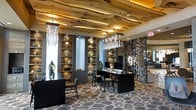 This single-floor space makes a striking impression through its thoughtful combination of natural and contemporary design elements. The ceiling is a standout feature, showcasing raw-edge wood beams with warm underlighting that adds an organic, rustic character juxtaposed against the otherwise modern interior. Suspended beneath are elegant crystal pendant chandeliers that cast a refined, ambient glow across the space.

Built-in shelving units line the walls, finished in a warm wood tone and set against dark grey surrounds, displaying decorative vessels, vases, and sculptural objects that add visual interest and curated character. A sleek dark workstation or reception desk sits centrally, equipped with monitors and task seating, suggesting a functional yet design-forward purpose.

A decorative laser-cut metal screen panel serves as a partial room divider, featuring a bold circular pattern that adds depth and visual texture. The floor is covered in a large-scale patterned carpet with organic, stone-like motifs in neutral tones with earthy green accents, anchoring the room's aesthetic. Overall, the space balances warmth, sophistication, and functionality with a cohesive and well-appointed design.