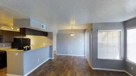 This single-story space in Lancaster offers a thoughtfully designed open layout that promotes a natural flow between living and dining areas. Rich, dark hardwood-style flooring runs throughout, adding warmth and a sense of cohesion to the interior. The dining area is anchored by an elegant pendant light fixture that draws the eye upward to a textured ceiling, creating a refined focal point in the room. A window fitted with blinds invites natural light into the space, brightening the grey-toned walls that lend a modern, neutral aesthetic throughout. The kitchen is connected to the main living area via a convenient pass-through counter with a light stone-toned surface, making the space feel open and connected while maintaining a functional separation. Dark cabinetry above the pass-through adds contrast and a bold design element. Ventilation is well-considered with a ceiling vent positioned for efficient airflow. The overall ambiance is clean and contemporary, with a neutral color palette and quality finishes that create a versatile environment suitable for a variety of uses. The single-floor layout ensures ease of movement and accessibility throughout the entire space.