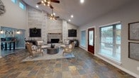 This thoughtfully designed single-floor commercial space blends rustic charm with modern functionality, creating a warm and welcoming atmosphere throughout. The centerpiece of the main lounge area is a soaring, floor-to-ceiling shiplap accent wall framing a decorative fireplace adorned with intricate patterned tile surround and a solid wood mantel. Vaulted ceilings amplify the sense of openness, while two ceiling fans provide both style and comfort. Multipoint recessed lighting and large windows allow abundant natural light to fill the space. Rich, multi-toned slate tile flooring flows seamlessly throughout, grounding the interior with an earthy, textural quality. The lounge is furnished with natural wood-framed chairs and a round metal coffee table, arranged to encourage conversation. A dedicated workspace transitions naturally from the lounge, featuring a substantial dark wood desk set against deep gray-painted walls, offering a focused yet cohesive professional environment. A compact kitchenette serves the space with dark cabinetry, open pipe shelving, a full-sized refrigerator, and countertop appliances — practical amenities for daily use. A stained glass-accented front entry door adds an artisanal touch to the welcoming entrance. Overall, the space exudes a refined farmhouse-industrial aesthetic ideal for a client-facing professional or service-oriented business.