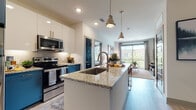 This thoughtfully designed single-floor residence combines contemporary style with functional living. The open-concept kitchen is a standout feature, boasting two-tone cabinetry in crisp white and bold teal, granite countertops, a large central island with an undermount sink, stainless steel appliances, and a subway tile backsplash — all illuminated by recessed lighting and stylish pendant fixtures. The kitchen flows seamlessly into the living area, where large sliding glass doors open to a private balcony, flooding the space with natural light and offering pleasant outdoor views of surrounding greenery.

The living room is welcoming and well-proportioned, featuring warm wood-toned flooring and a layout that encourages both relaxation and entertaining. Two bedrooms provide comfortable retreats; one features a striking exposed brick accent wall, plush carpeting, large windows, and a ceiling fan, while the second offers a bright, serene atmosphere with generous natural light. Both bathrooms are finished with granite-topped teal vanities, light wood-style flooring, and walk-in closets with built-in shelving providing ample organization. The bathrooms also include glass-enclosed showers and well-lit vanity mirrors, reflecting a high level of attention to detail throughout the residence.