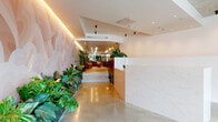 This thoughtfully designed amenity floor makes a striking first impression with a grand reception area finished in polished concrete floors and a statement marble-clad desk. Lush tropical greenery lines the entry corridor alongside a softly lit, curved feature wall adorned with an abstract pastel mural, guiding visitors deeper into the space.

The lounge area is a standout, featuring plush velvet seating, a grand piano, and decorative guitars mounted on the wall — all centered around expansive floor-to-ceiling glass doors that open to an outdoor terrace furnished for relaxation. An ornate embossed ceiling panel and warm ambient lighting add a layer of refined character to this social hub.

A dedicated co-working area offers long natural timber tables paired with classic dark chairs and brass mushroom table lamps, set within a glass-walled room that maintains visual connection to surrounding greenery and lounge spaces — ideal for focused work or collaboration.

A media lounge provides a more intimate setting, complete with a large wall-mounted TV, a marble-topped island, warm timber cabinetry, and built-in shelving, creating a comfortable retreat for informal gatherings.

The fitness center rounds out the offering with a comprehensive suite of cardio machines, free weights, and strength equipment set on light timber flooring beneath an exposed concrete ceiling — functional, spacious, and well-appointed.