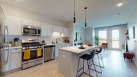 This single-floor residence offers a thoughtfully designed open-concept layout that seamlessly connects the kitchen, dining, and living areas. The kitchen is a standout feature, showcasing crisp white cabinetry, a herringbone tile backsplash, stainless steel appliances including a gas range, dishwasher, and microwave, as well as a generous island with a white quartz countertop and pendant lighting overhead. Light wood-style flooring runs throughout the main living areas, enhancing the bright and airy atmosphere.

The living room is filled with natural light from large windows and a glass door that opens to a private balcony, offering outdoor views. An exposed ductwork detail adds an industrial accent to the otherwise clean, modern aesthetic. The space is further complemented by open wall shelving and ample room for comfortable furnishings.

The residence includes two bedrooms, each carpeted and bathed in natural light from wide windows. One bedroom features a ceiling fan, open outdoor views, and space for a desk, while the other provides a cozy, well-proportioned retreat. The bathroom is finished with a gray vanity, white countertop, large mirror, and modern matte black fixtures, with access to an adjacent laundry area for added convenience.