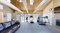 This single-floor community amenity space combines fitness and social gathering functions under one generously proportioned roof. The architecture is immediately striking, defined by soaring vaulted ceilings clad in warm natural wood planking and supported by exposed white-painted beams. A central skylight draws abundant natural light deep into the interior, complementing the large windows that line the perimeter walls and offer views of the surrounding outdoor area.

The fitness zone is well-equipped, featuring treadmills, an elliptical trainer, a stationary bike, and a cable weight machine, all positioned on plush grey carpeting. A brick fireplace with a wall-mounted flat-screen television above it serves as a focal point, adding a welcoming residential character to the exercise area. Yellow accent walls near the ceiling add a subtle pop of color throughout the space.

A lounge and reading area provides comfortable leather seating alongside a round table with chairs and a bookshelf stocked with reading material, creating a relaxed social environment. A compact kitchen is outfitted with white cabinetry, full-size refrigerator, dishwasher, microwave, and additional countertop appliances, making the space well-suited for gatherings. A small workstation and utility areas complete this versatile, multi-functional facility.