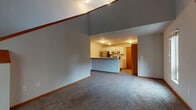 Located at 1202 McKenna Blvd in Madison, Wisconsin, this two-floor residence offers a functional and well-organized layout across both levels. The main level features an open-concept living and dining area with plush carpeting, neutral gray walls, and soaring vaulted ceilings that lend a sense of expansive vertical space. A kitchen island with a breakfast bar connects the living area to the kitchen, which is equipped with white cabinetry, stainless steel appliances including a refrigerator and electric range, and light-toned countertops. A staircase with wood railings leads to the upper level, where carpeted bedrooms offer a quiet retreat. At least one bedroom includes a sliding glass door that opens to an outdoor deck, providing views of surrounding greenery. A full bathroom on the main level features a tub-shower combination, a white vanity, and wood-look flooring. The entry door, with its warm wood finish, adds a classic touch to the space. Throughout both floors, warm wood baseboards and trim provide a cohesive design element, while ample windows allow natural light to flow throughout the home.