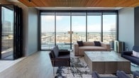 This refined single-floor high-rise space masterfully blends contemporary design with luxurious functionality. A striking architectural feature throughout is the warm wood slat ceiling, which adds texture and warmth while creating a cohesive visual identity across all areas.

The expansive living and lounge area is anchored by dramatic floor-to-ceiling glass curtain walls that frame breathtaking panoramic city views. Natural light floods the space, illuminating a carefully curated seating arrangement that includes a plush channeled sofa, sleek armchairs with brass-accented legs, and a marble-top coffee table set atop a layered area rug over wide-plank hardwood flooring.

The gourmet kitchen is a standout feature, offering professional-grade stainless steel appliances, including a commercial-style range and built-in ovens. Custom wood cabinetry lines the walls alongside a crisp white marble backsplash, with large-format stone tile flooring grounding the space. A substantial island provides both workspace and visual separation.

Adjacent to the kitchen, a sophisticated bar area features gold geometric pendant lighting, a marble backsplash, and backlit open shelving set against exposed brick—adding an industrial contrast to the otherwise polished aesthetic. Brushed gold bar stools complete the refined yet inviting atmosphere, making this space ideal for both entertaining and relaxed everyday use.
