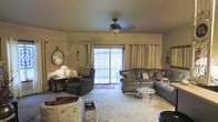 This single-floor space centers around a warmly appointed living room designed for both comfort and functionality. The room is anchored by a large sliding glass door framed by floor-length grayish-beige grommet curtains, which allow generous natural light to fill the space while offering views of an open outdoor landscape. A ceiling fan with a light fixture is centrally mounted, providing both ventilation and ambient lighting. The seating arrangement includes a large sectional sofa with decorative pillows and a matching recliner chair, both upholstered in a neutral gray-brown fabric. A round glass-top coffee table with ornate metal legs serves as a central accent piece. The flooring is covered by a large area rug featuring a light floral pattern. Along the walls, thoughtful decorative touches include an oversized vintage-style wall clock and wall-mounted accents that add character to the neutral-toned walls. A small side table with a lamp and personal items occupies one corner, alongside what appears to be a pet crate, suggesting a pet-friendly environment. The overall ambiance is relaxed and lived-in, with a welcoming, homey atmosphere.