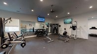 This fitness center is thoughtfully organized into distinct workout zones, offering a comprehensive range of equipment to support varied exercise routines. The space features durable rubber flooring throughout, providing safety and comfort underfoot, while crisp white walls and recessed lighting create a clean, functional atmosphere.

The strength training area is well-appointed with a full dumbbell rack spanning multiple weight increments, a cable machine system, a weight bench with barbell setup, and a power tower for bodyweight exercises. A large wall-mounted mirror spans one side of this zone, enhancing the sense of space and allowing users to monitor their form. Two wall-mounted flat-screen TVs are positioned for easy viewing during workouts, and a ceiling fan ensures comfortable air circulation.

The cardio zone features treadmills, elliptical trainers, and stationary bikes, complemented by an additional wall-mounted TV and a ceiling fan. Natural light filters in through windows fitted with roller shades.

A dedicated functional training area houses stability balls, medicine balls, resistance bands, a back extension bench, and additional free weights. Built-in cabinetry provides storage. Rowing machines occupy a separate section of the space, accompanied by another mounted television. The overall layout promotes an efficient, distraction-free training environment.