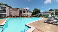 This outdoor amenity area centers around a generously sized swimming pool with clear blue water, framed by a broad concrete pool deck and classic brick-paved pathways that define the courtyard space. The pool area is well-suited for leisure and recreation, offering lounge seating and a pergola structure that provides a shaded gathering spot adjacent to the water.

Mature trees, including crape myrtles and magnolias, are planted throughout the courtyard, offering natural shade and a sense of established landscaping. Brick accent walls and low fencing define the perimeter of the space, creating distinct zones while maintaining open sightlines across the grounds. Trash receptacles and other functional amenities are thoughtfully integrated into the design.

The surrounding multi-story residential buildings feature balconies, neutral siding, and pitched rooflines that give the community a traditional architectural character. Access to the adjacent building is provided through white-framed glass doors, which open directly onto the brick patio. Landscaped shrubs and groundcover line the walkways, contributing to the overall well-maintained and inviting ambiance of the outdoor common area.