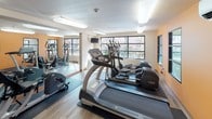 Located at 95 Burnett Ave S in Renton, Washington, this single-floor fitness center is designed to support a well-rounded workout routine. The space is outfitted with a variety of cardio equipment, including treadmills and elliptical trainers, as well as a multi-station cable strength machine, a rowing machine, and a stationary bike — offering options for both cardiovascular and resistance training.

The room features wood-look luxury vinyl flooring throughout, with protective rubber mats positioned beneath the equipment. A large wall mirror along one side of the room visually expands the space and assists with form during exercise. Vibrant orange accent walls add an energetic ambiance, while framed motivational artwork contributes to the motivating atmosphere.

Natural light fills the room through a series of large, multi-pane windows that frame views of the surrounding neighborhood, including mature cherry blossom trees. A wall-mounted ductless mini-split air conditioning unit ensures year-round climate comfort. Overhead fluorescent lighting provides consistent illumination throughout. The space also includes posted instructional and informational materials near the entrance, making it accessible and user-friendly for individuals of all fitness levels.