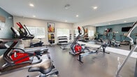Located at 1459 KLO Road in Kelowna, British Columbia, this dedicated fitness centre is a thoughtfully outfitted exercise facility designed to support a full range of workout routines. The space features durable rubber flooring throughout, recessed ceiling lighting, and multiple windows that allow natural light to fill the room, creating a bright and energizing environment.

The cardio zone is well-appointed with treadmills, rowing machines, a stationary bike, and an elliptical trainer, each positioned in front of wall-mounted flat-screen televisions for an engaging workout experience. A bold teal accent wall anchors this section of the room, adding a modern design touch.

The strength training area includes a multi-function cable machine, a weight bench, a full rack of dumbbells in graduated sizes, medicine balls, and additional free weight accessories stored on dedicated racks. A large wall mirror expands the sense of space and allows users to monitor their form during exercise.

Vibrant, colourful artwork adorns the white walls, contributing to an upbeat and motivating atmosphere. A storage unit provides space for smaller accessories and personal items. The facility is also climate-controlled for year-round comfort. Overall, this fitness centre delivers a comprehensive and well-organized workout environment suitable for residents of all fitness levels.