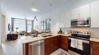 This single-story 1-bedroom, 1-bathroom residence offers a thoughtfully designed open-concept layout that seamlessly connects the kitchen, dining, and living areas. The kitchen is a standout feature, equipped with stainless steel appliances—including a range, microwave, and dishwasher—complemented by rich walnut-toned lower cabinetry, crisp white upper cabinets, a subway tile backsplash, and light quartz countertops. Pendant lighting adds a modern touch above the kitchen peninsula, which also functions as a convenient breakfast bar.

The adjacent dining area is defined by light wood-look flooring and tall walls adorned with large-scale framed artwork. The living room receives generous natural light through oversized windows dressed with patterned drapes, and features a ceiling fan and ample space for comfortable furnishings.

The bedroom is carpeted and well-lit by a large window with outdoor views, offering a tranquil retreat. A second bedroom variation showcases a bold geometric accent wall for added character. The bathroom is finished with large-format tile flooring, a glass-enclosed shower with tile surround, and access to a walk-in closet area, completing this stylish and functional residence.