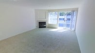 This single-floor residence features a spacious, open living area designed with a clean, minimalist aesthetic. The room is finished with soft, plush wall-to-wall carpeting in a neutral tone that complements the crisp white walls, creating a bright and airy atmosphere throughout. A centrally positioned fireplace with a classic black surround serves as a natural focal point, adding warmth and character to the space.

Natural light fills the room generously, courtesy of a large picture window positioned alongside a sliding glass door that opens to an outdoor area. The outdoor views reveal a balcony-style setting with glimpses of surrounding greenery and neighboring structures. The sliding door arrangement not only maximizes light penetration but also provides convenient indoor-outdoor access, enhancing the sense of openness.

The layout is notably open and versatile, offering ample room for a variety of furniture arrangements. The neutral color palette of whites and beiges throughout the walls, trim, and flooring ensures the space can adapt easily to a range of interior styles. Overall, the living area conveys a comfortable, well-lit environment with practical yet appealing architectural details.