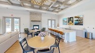 Located at 542 Gateway Centre Pkwy in North Chesterfield, VA, this single-floor community clubroom offers a well-appointed, open-plan gathering space with a thoughtful blend of comfort and sophistication. The room is anchored by a striking coffered ceiling adorned with a contemporary sputnik chandelier, lending architectural character and visual height throughout. Light hardwood flooring runs seamlessly underfoot, complementing the bright, white walls and creating a warm, welcoming atmosphere.

A floor-to-ceiling stacked stone fireplace with a wood mantel serves as a dramatic focal point in the dining and lounge area, where round tables and mixed seating invite casual gatherings. Adjacent to this is a substantial white island with a light quartz countertop and bar-height seating, ideal for socializing or dining. The island is complemented by a built-in sink and cabinetry, enhancing the space's functionality.

The lounge zone features rich cognac leather seating arranged around a wall-mounted flat-screen TV and a textured credenza, creating a relaxed entertainment nook. Large windows flood the entry area with natural light and offer views of the surrounding outdoor environment, while glass double doors provide direct access to an exterior pool area. The overall ambiance is modern, polished, and community-focused.