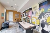 Located at 3400 Davie Rd in Davie, Florida, this dynamic hallway serves as a lively transitional and recreational space within the building. The standout feature is a sweeping, large-scale mural inspired by Jean-Michel Basquiat's iconic neo-expressionist style, spanning an entire wall with bold colors, expressive figures, and striking text elements that instantly command attention and set an energetic tone throughout the space.

The hallway is thoughtfully outfitted for recreation, housing a full-size Brunswick air hockey table as its centerpiece, alongside a classic arcade game cabinet positioned along the adjacent wall. A cozy bistro-style high-top table with chairs provides a casual seating nook, complemented by colorful abstract artwork hung on a warm amber accent wall nearby.

The flooring features a patterned stone or tile design in muted grey tones, adding texture and visual interest underfoot. Recessed ceiling lighting provides even, well-distributed illumination throughout. Floor-to-ceiling glass doors and windows flood the space with natural light and offer direct views and access to an outdoor pool courtyard lined with palm trees, seamlessly blending indoor and outdoor living. A glass-paned door connects the hallway to the building's main lobby, ensuring smooth circulation and an open, connected feel.