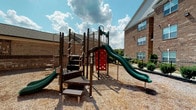 This dynamic community amenity space combines outdoor recreation with an elegantly designed indoor clubhouse. At its heart is a multi-level playground structure set on a wood chip surface, featuring green slides, a red climbing wall, staircases, and multiple elevated platforms — all framed by lush greenery and brick residential buildings under open skies.

The adjacent clubhouse is a standout gathering space anchored by floor-to-ceiling glass walls that flood the interior with natural light and offer direct views of the resort-style pool and outdoor lounge area. The polished concrete floors throughout lend a sleek, modern feel to the space.

Inside, the clubhouse offers a welcoming lounge area with a large sectional sofa, teal accent chairs, and a stone fireplace topped with a wall-mounted television — ideal for relaxed socializing. A vaulted ceiling with decorative pendant and chandelier lighting enhances the sense of openness. The dining and café area features high-top tables and chairs in teal and yellow tones, complemented by a teal accent wall and vibrant artwork. A small bar or service counter adds a practical touch. The seamless indoor-outdoor connection, contemporary color palette, and thoughtfully curated furnishings create a lively yet comfortable community environment.
