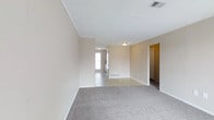 This single-floor space offers a straightforward and functional layout with a calm, neutral aesthetic throughout. The living area is generously proportioned, featuring soft gray carpeting underfoot and walls painted in a warm beige tone that creates a welcoming, understated atmosphere. Crown molding lines the ceiling, adding a subtle architectural detail that elevates the overall finish of the room. Ceiling-mounted light fixtures provide steady illumination, while a visible HVAC vent ensures year-round climate comfort. The open sightline connects the carpeted living area to an adjoining space with hard flooring, suggesting a natural transition toward a kitchen or dining area. A window at the far end allows natural light to filter into the interior, brightening the connected spaces. Doorways branch off to additional rooms, hinting at a well-organized floor plan. White baseboards trim the perimeter of each room, complementing the clean ceiling and walls. The overall ambiance is neat, move-in ready, and well-maintained, making it a blank canvas well-suited to a variety of furniture arrangements and personal styles.