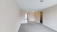 This single-floor space offers a straightforward and functional layout with a calm, neutral aesthetic throughout. The living area is generously proportioned, featuring soft gray carpeting underfoot and walls painted in a warm beige tone that creates a welcoming, understated atmosphere. Crown molding lines the ceiling, adding a subtle architectural detail that elevates the overall finish of the room. Ceiling-mounted light fixtures provide steady illumination, while a visible HVAC vent ensures year-round climate comfort. The open sightline connects the carpeted living area to an adjoining space with hard flooring, suggesting a natural transition toward a kitchen or dining area. A window at the far end allows natural light to filter into the interior, brightening the connected spaces. Doorways branch off to additional rooms, hinting at a well-organized floor plan. White baseboards trim the perimeter of each room, complementing the clean ceiling and walls. The overall ambiance is neat, move-in ready, and well-maintained, making it a blank canvas well-suited to a variety of furniture arrangements and personal styles.