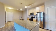Located at 9101 Stony Point Pkwy in Richmond, Virginia, this single-floor residence offers a well-designed, open-concept layout with modern finishes throughout. The kitchen serves as a central hub of the home, anchored by a large granite-topped island with an integrated sink and pendant lighting overhead. Light maple cabinetry lines the walls, complemented by a full suite of stainless steel appliances including a refrigerator, electric range, and built-in microwave. Wood-look flooring extends throughout the main living areas, creating a cohesive and easy-to-maintain aesthetic. The open floor plan seamlessly connects the kitchen to the living area, which is bright and spacious with neutral-toned walls. The bedroom features plush carpeting, a generous closet with wire shelving, and a window that allows natural light to fill the room. The bathroom is finished with granite countertops matching the kitchen, warm wood cabinetry, a large mirror, and a tiled tub-shower combination. A dedicated laundry area houses a full-size top-loading washer and dryer with additional overhead shelving for storage. The overall design is clean, contemporary, and functional, making it a comfortable and practical living space in the Stony Point area of Richmond.